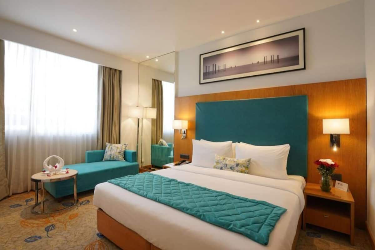 Quarto de hotel com uma cama grande, um sofá, uma mesinha, uma mesa de cabeceira, uma luminária, um espelho e um quadro na parede.
