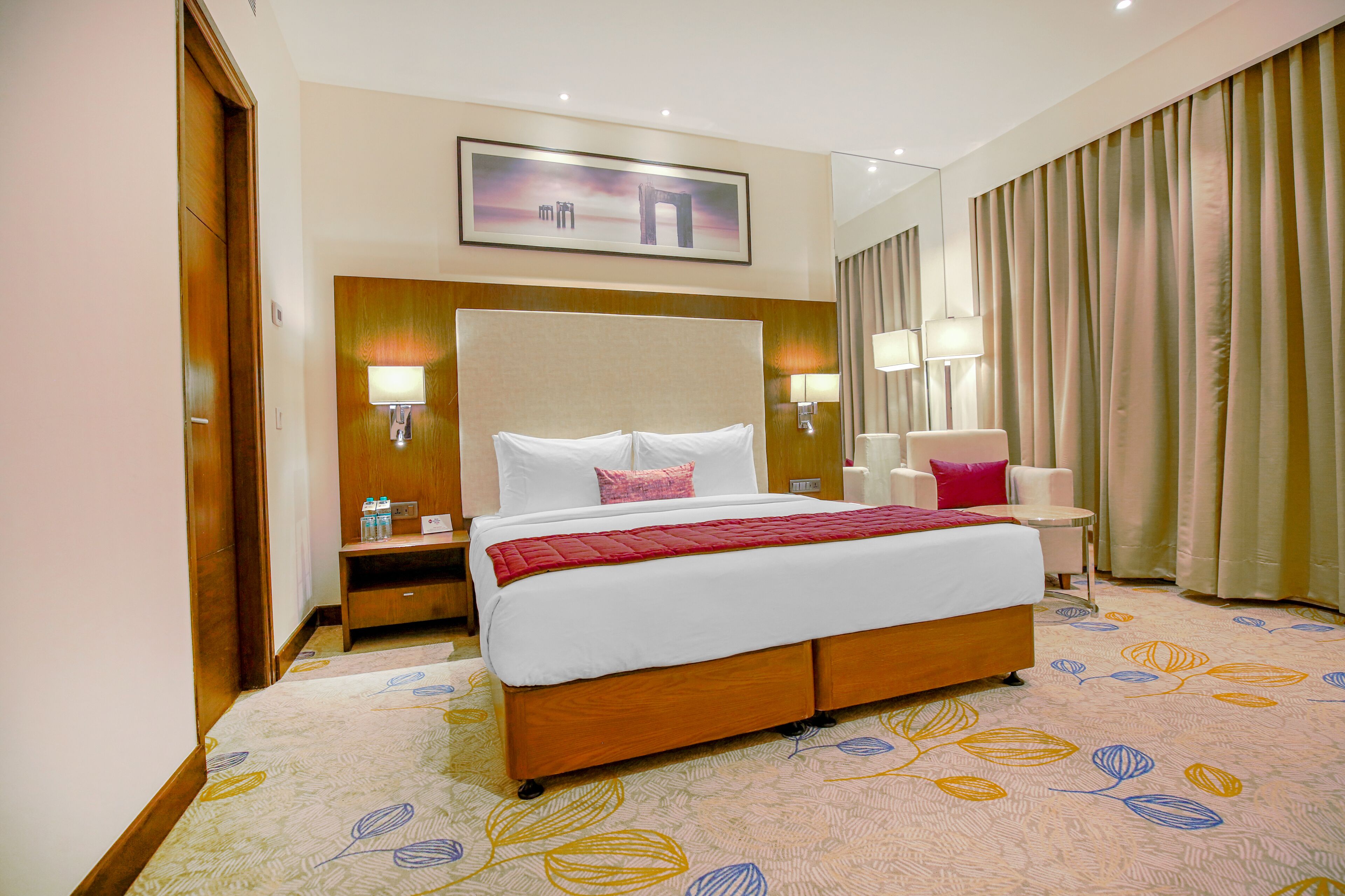 Une chambre d’hôtel avec un grand lit, des tables de chevet, une petite table, une chaise, un bureau et une grande fenêtre avec des rideaux.