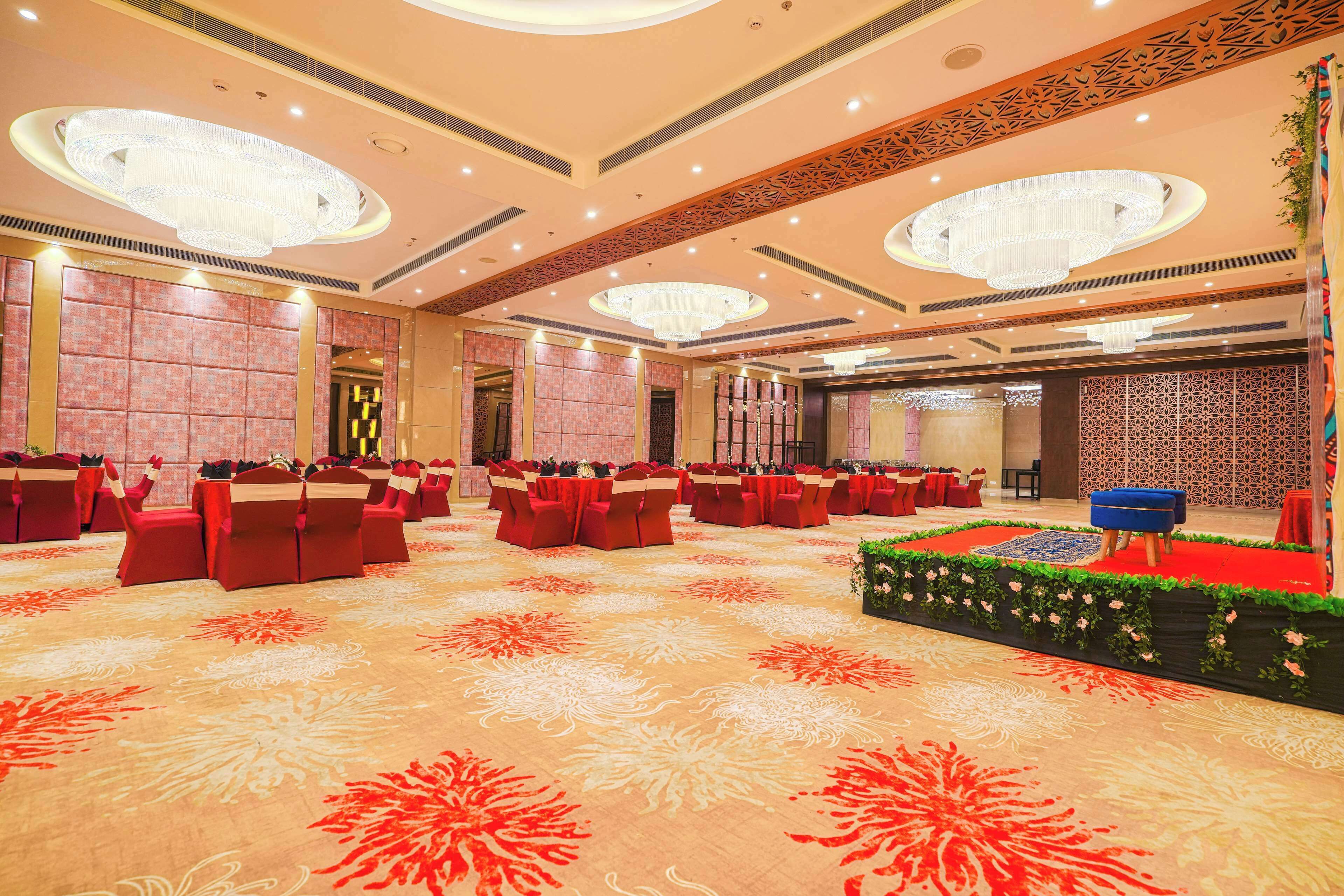 A spacious banquet hall with multiple round tables, red chairs, and a central stage with a blue sofa.