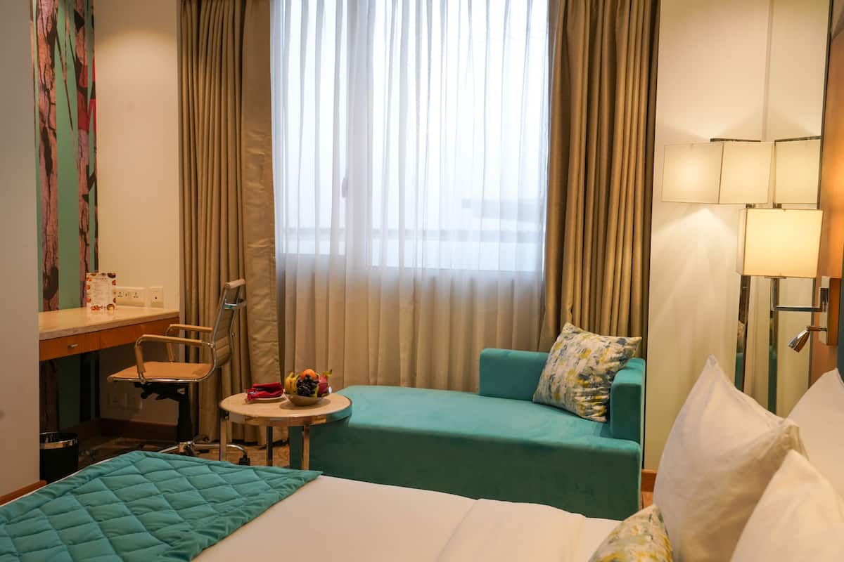 Quarto de hotel com cama, uma pequena mesa com frutas, uma poltrona azul-esverdeada e uma escrivaninha com cadeira.