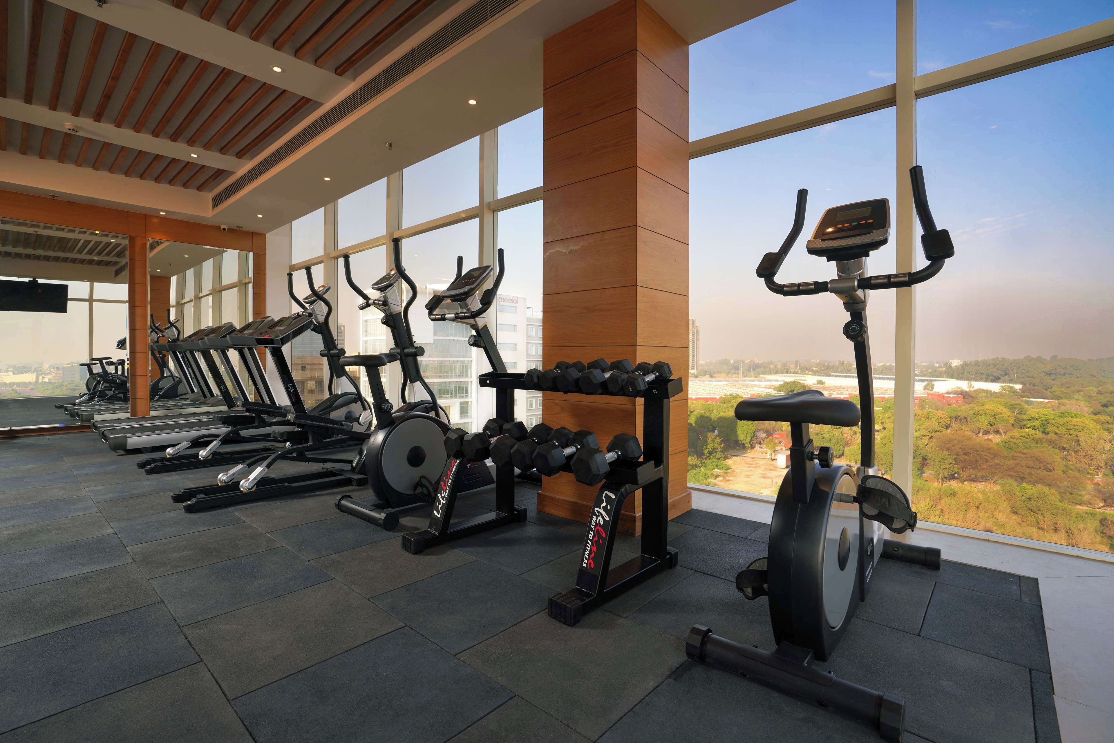A gym with treadmills, elliptical machines, and a stationary bike, with large windows offering a view of the city.