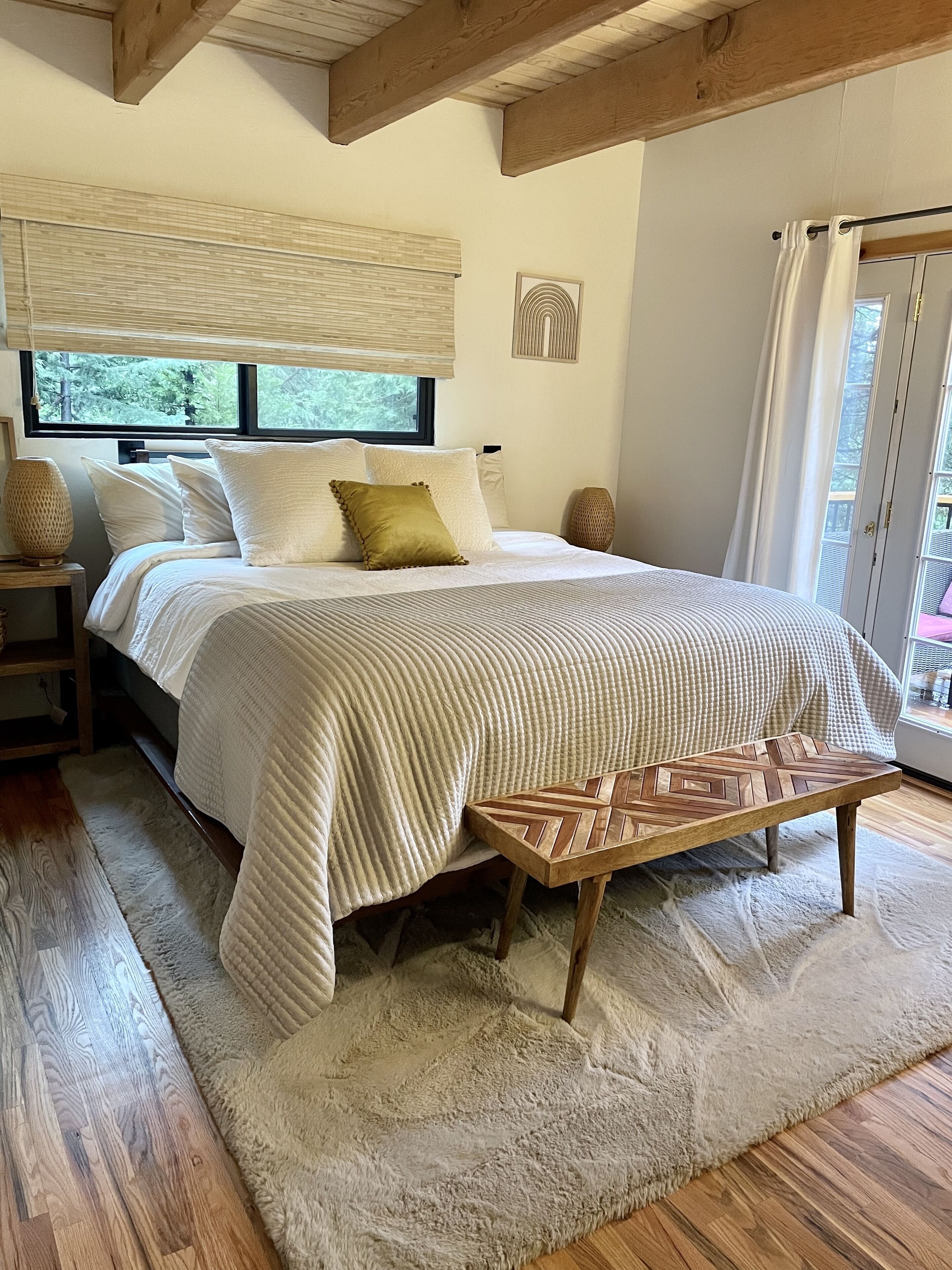 A bedroom with a large bed, a wooden bench, a fluffy rug, and a window with blinds.