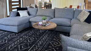 A spacious living room with a large gray sectional sofa, a round coffee table, and a black and white patterned rug. A staircase leads to an upper level with a wooden railing.