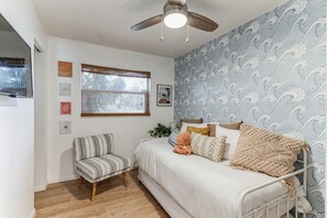 Un dormitorio con cama, una silla, ventilador de techo y ventana con persianas.