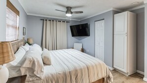 Ein Schlafzimmer mit einem Bett, einem Deckenventilator, einem an der Wand montierten Fernseher, einem Kleiderschrank und einem Fenster mit Vorhängen.