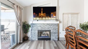 A living room with a flat-screen TV mounted above a stone fireplace, a dining table with chairs, and a view of the Eiffel Tower at night.