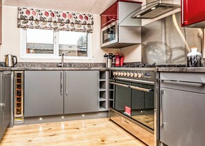 A kitchen with grey cabinets, a stainless steel range hood, a built-in oven, and a window with a floral valance. - 3 bedroom accommodation in Carnforth (Carnforth)