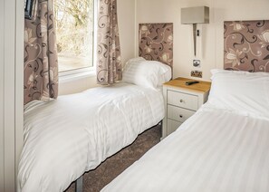 A bedroom with two beds, a bedside table with a lamp, and a window with curtains. - 3 bedroom accommodation in Carnforth (Carnforth)