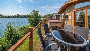 A deck with outdoor furniture overlooking a lake, a hot tub, and a wooden building with large windows. - 3 bedroom accommodation in Carnforth (Carnforth)