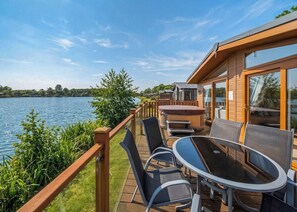 A deck with outdoor furniture overlooking a lake, a hot tub, and a wooden building with large windows. - 3 bedroom accommodation in Carnforth (Carnforth)