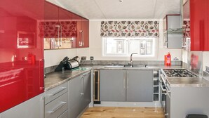 A modern kitchen with grey cabinets, a red accent wall, a wooden floor, and a window with a floral curtain.