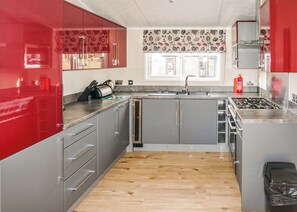 A modern kitchen with grey cabinets, a red accent wall, a wooden floor, and a window with a floral curtain.