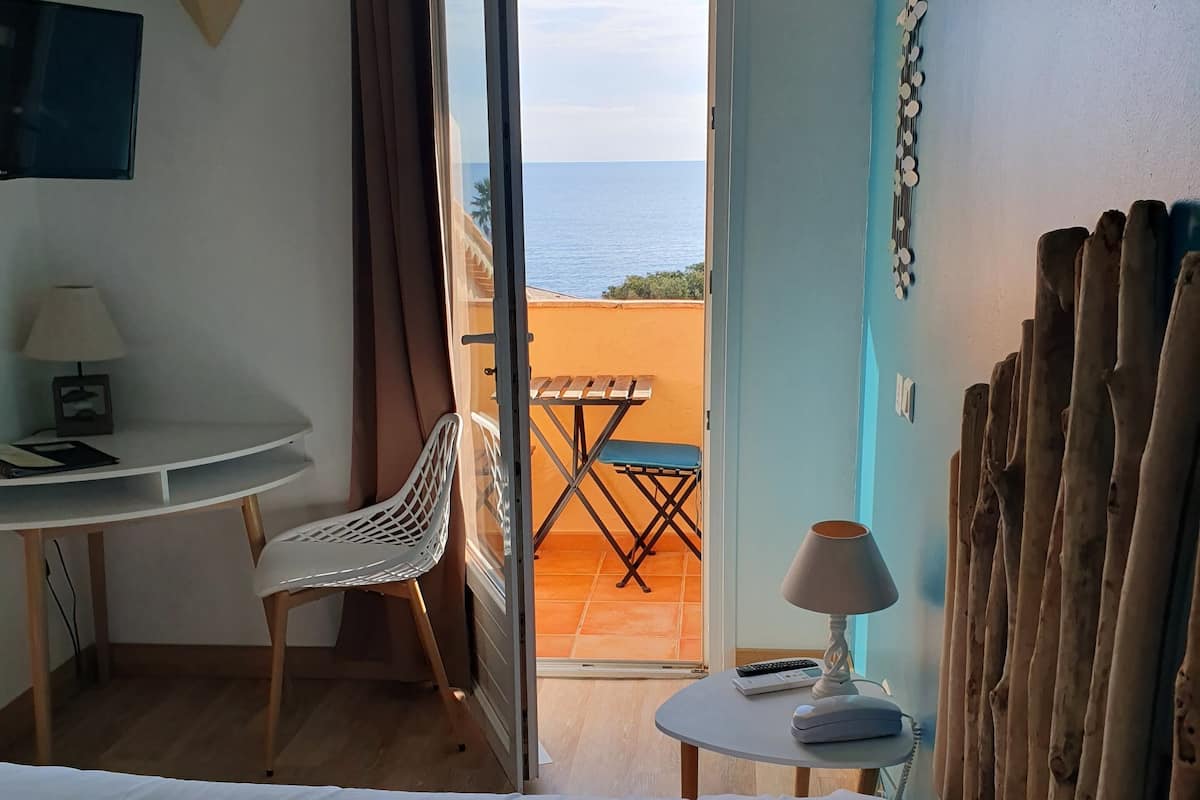 Ein Hotelzimmer mit Bett, Schreibtisch, Stuhl, kleinem Tisch mit Lampe und Meerblick durch eine offene Tür.