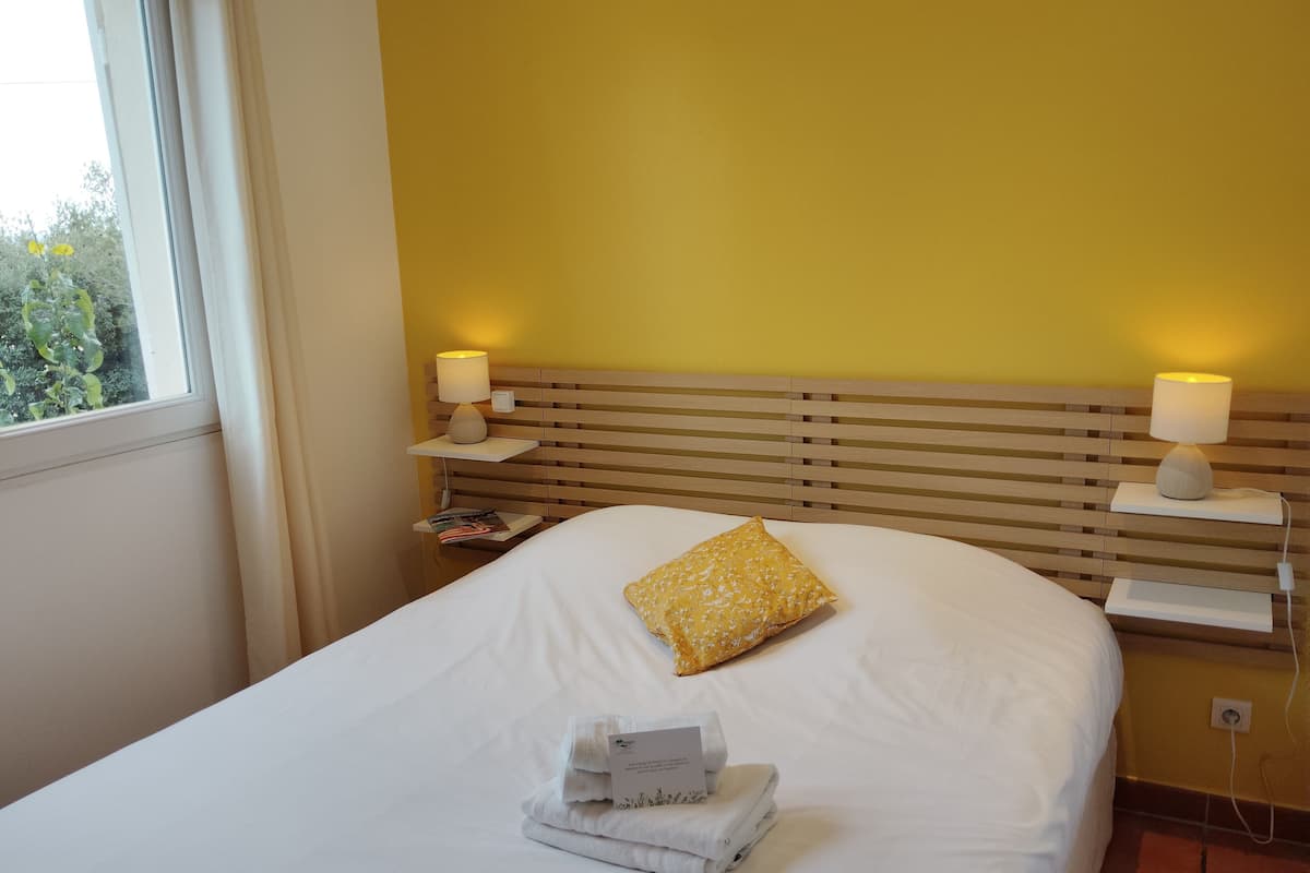 Ein ordentlich bezogenes Bett mit einem gelben Kissen, zwei Nachttischlampen, einem Nachttisch mit einer Zeitschrift und einem Fenster mit Vorhängen.