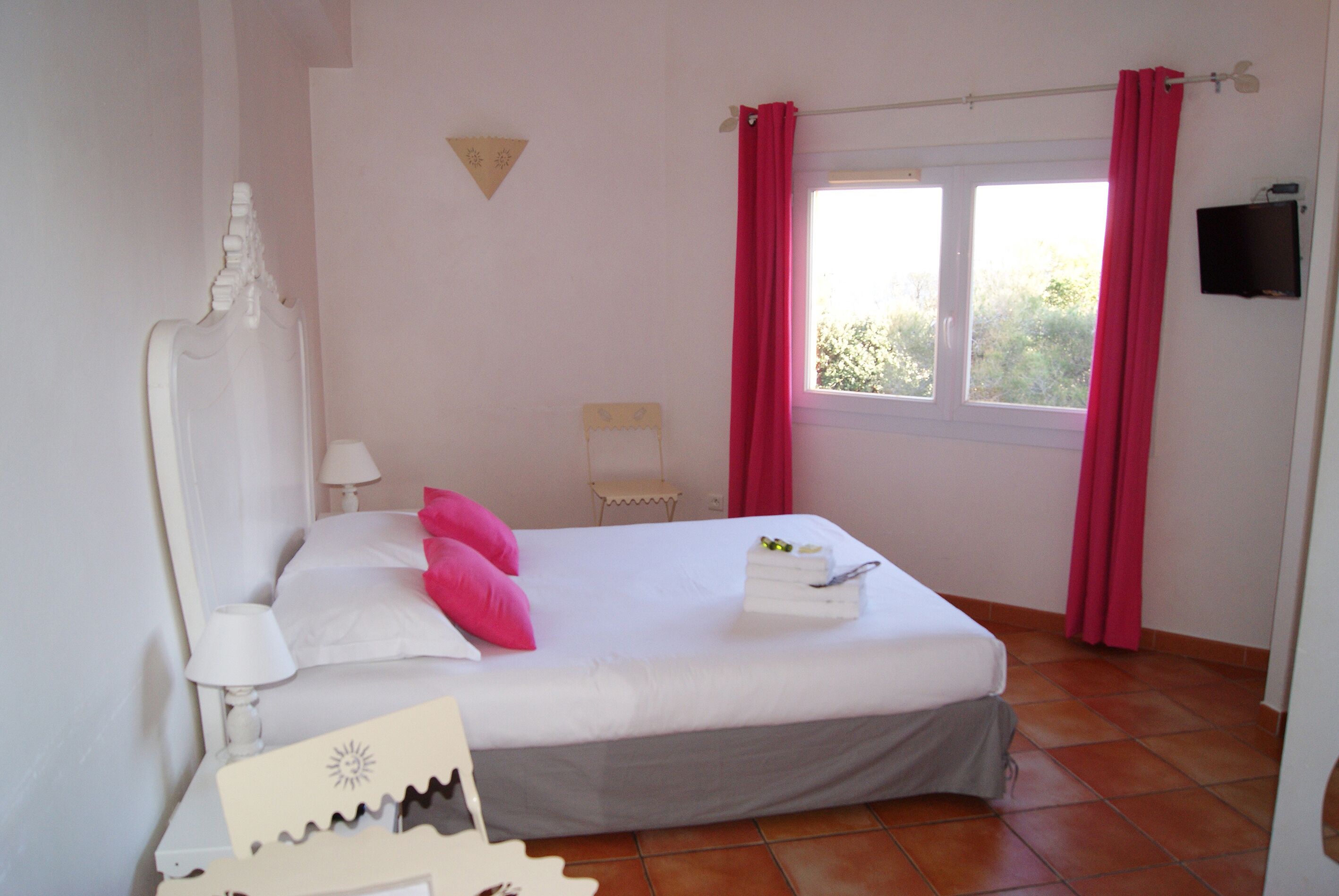 Un dormitorio con cama, almohadas, una mesita de noche, lámpara, ventana con cortinas y un televisor montado en la pared.