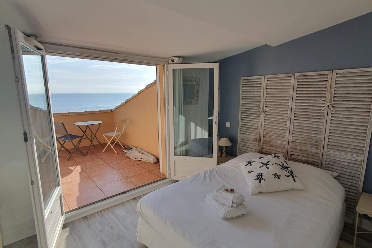 Ein Schlafzimmer mit Bett, einer Schiebetür zu einem Balkon mit Tisch und Stühlen sowie Blick aufs Meer.