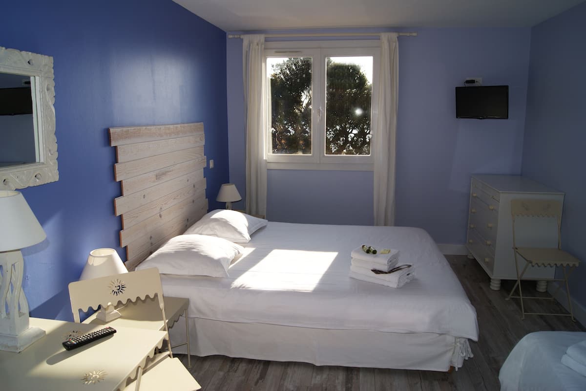 Ein Schlafzimmer mit Bett, Nachttisch, Kommode, Fernseher und einem Fenster mit Blick auf Bäume.