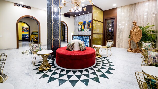 Een moderne lobby met een centraal rood, circulair zitgedeelte, goud- en witmeubilair, een grote kroonluchter en een marmeren vloer met een geometrisch patroon.