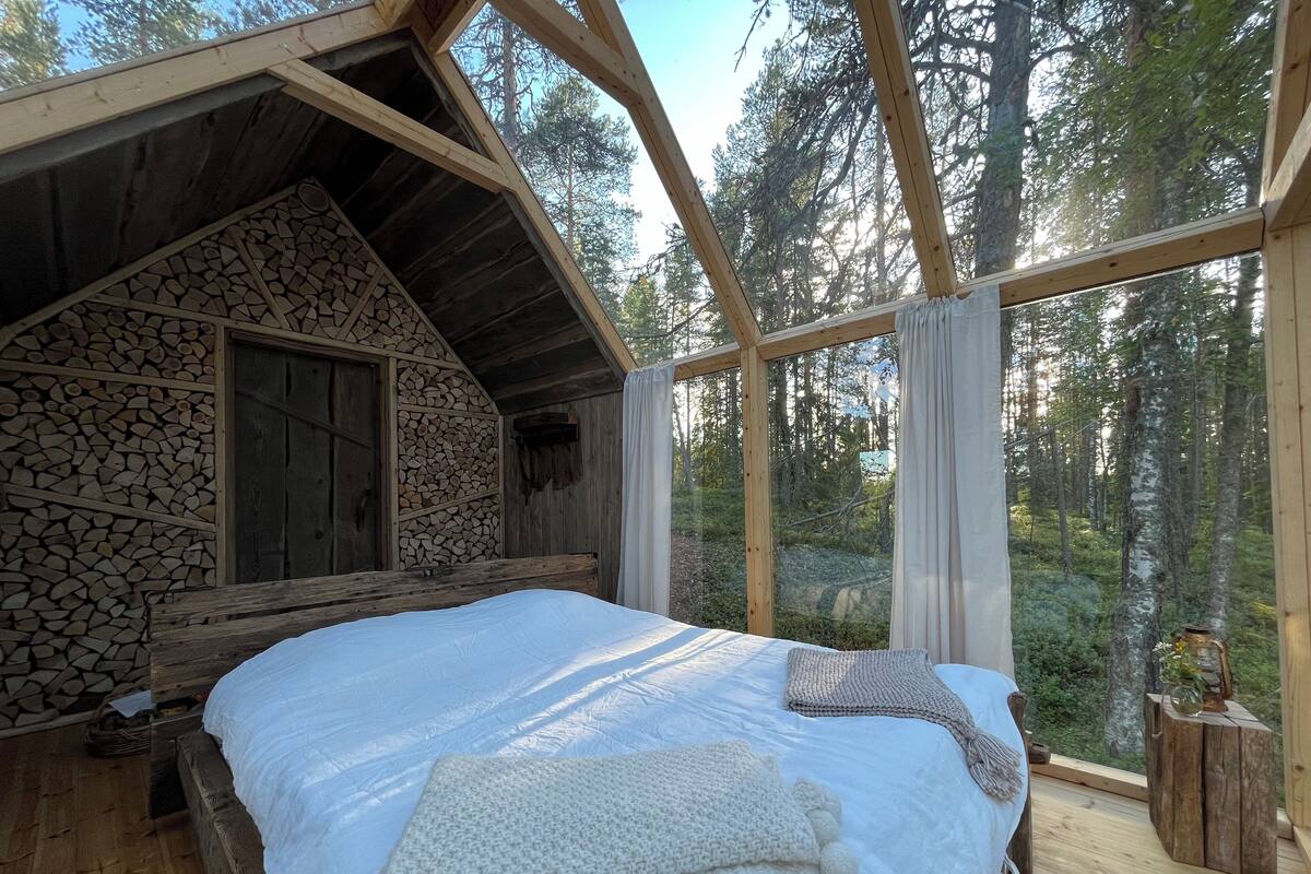 A cabin with a wooden interior, a bed with white bedding, and a view of the forest through large windows.