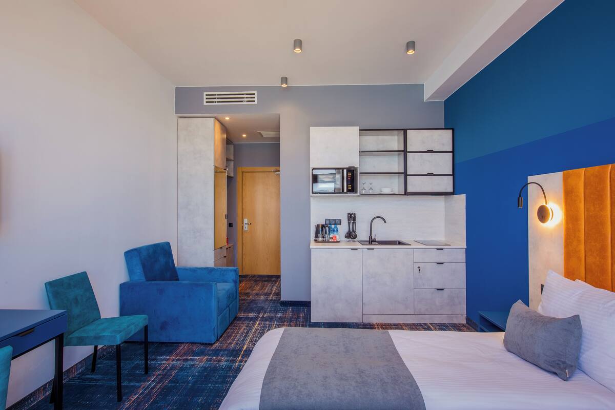 Une chambre d’hôtel moderne avec un grand lit, un coin salon avec des fauteuils bleus, une kitchenette et une salle de bain visible à travers une porte ouverte.