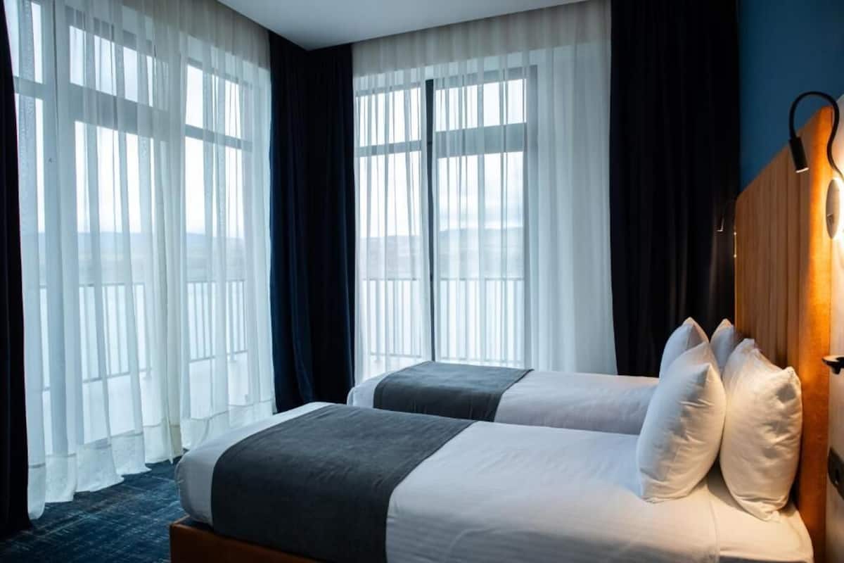 Une chambre d’hôtel avec deux lits, une tête de lit en bois, des rideaux légers et une vue sur la ville.