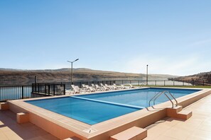 A rectangular swimming pool with clear blue water, surrounded by a black metal fence, and a series of white lounge chairs aligned along the pool's edge.