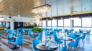A spacious dining area with blue upholstered chairs and round tables, large windows, and hanging chandeliers.
