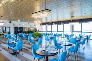 Een ruime eethoek met blauwe fauteuils en ronde tafels, grote ramen en hangende kroonluchters.