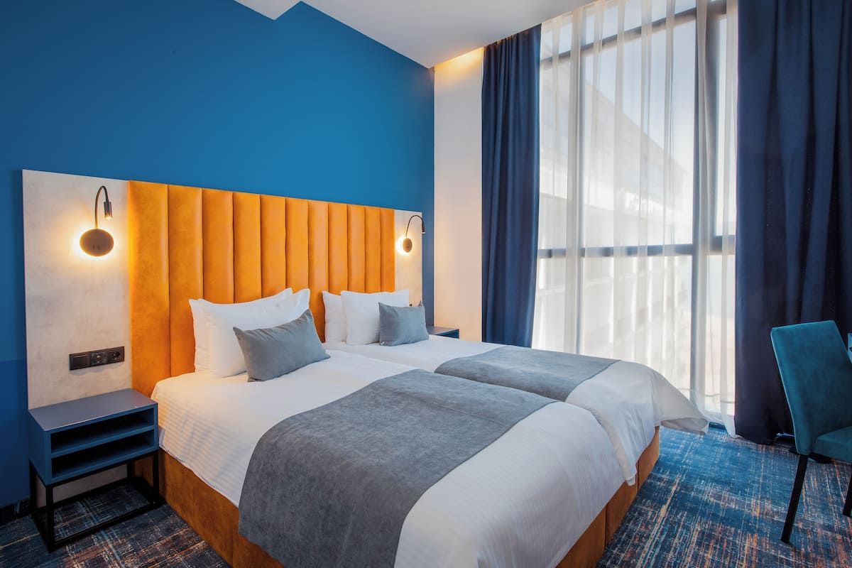 Une chambre d’hôtel avec un grand lit, deux tables de chevet, une petite table, une chaise et une grande fenêtre avec des rideaux.