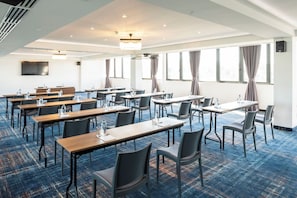 Une salle de conférence dotée de plusieurs tables et chaises, de grandes fenêtres avec des rideaux et d’un téléviseur à écran plat fixé au mur.