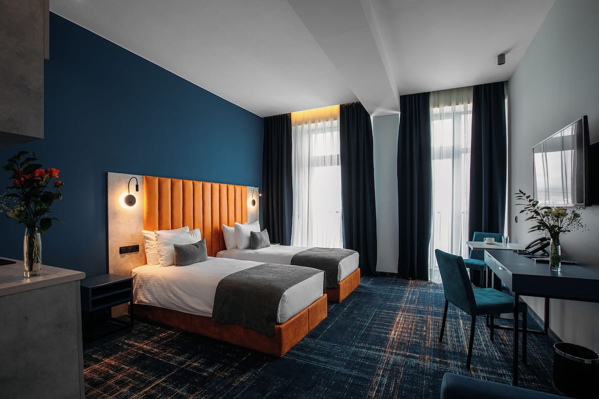 Une chambre d’hôtel avec deux lits, un bureau, une chaise et une grande fenêtre avec des rideaux.