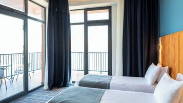 Ein Hotelzimmer mit zwei Betten, einem Balkon mit Tisch und Stühlen sowie großen Fenstern mit Vorhängen.