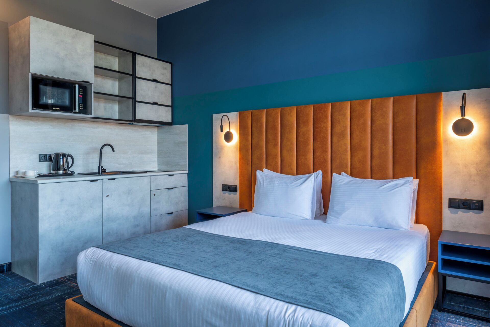 Una habitación de hotel moderna con cama, kitchenette e iluminación integrada en la cabecera.