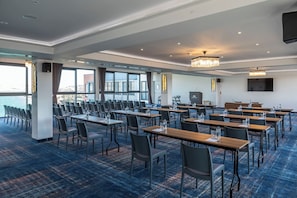 Une salle de conférence avec des rangées de tables et de chaises, de grandes fenêtres et un lustre.