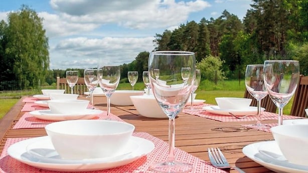 A wooden table set for a meal with white bowls and wine glasses, surrounded by a natural landscape.