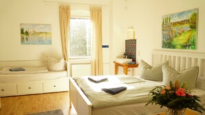 A bedroom with a bed, a bedside table with a lamp, a window with curtains, a painting on the wall, and a vase with flowers on a table.