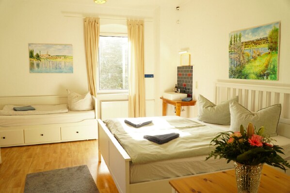 A bedroom with a bed, a bedside table with a lamp, a window with curtains, a painting on the wall, and a vase with flowers on a table.