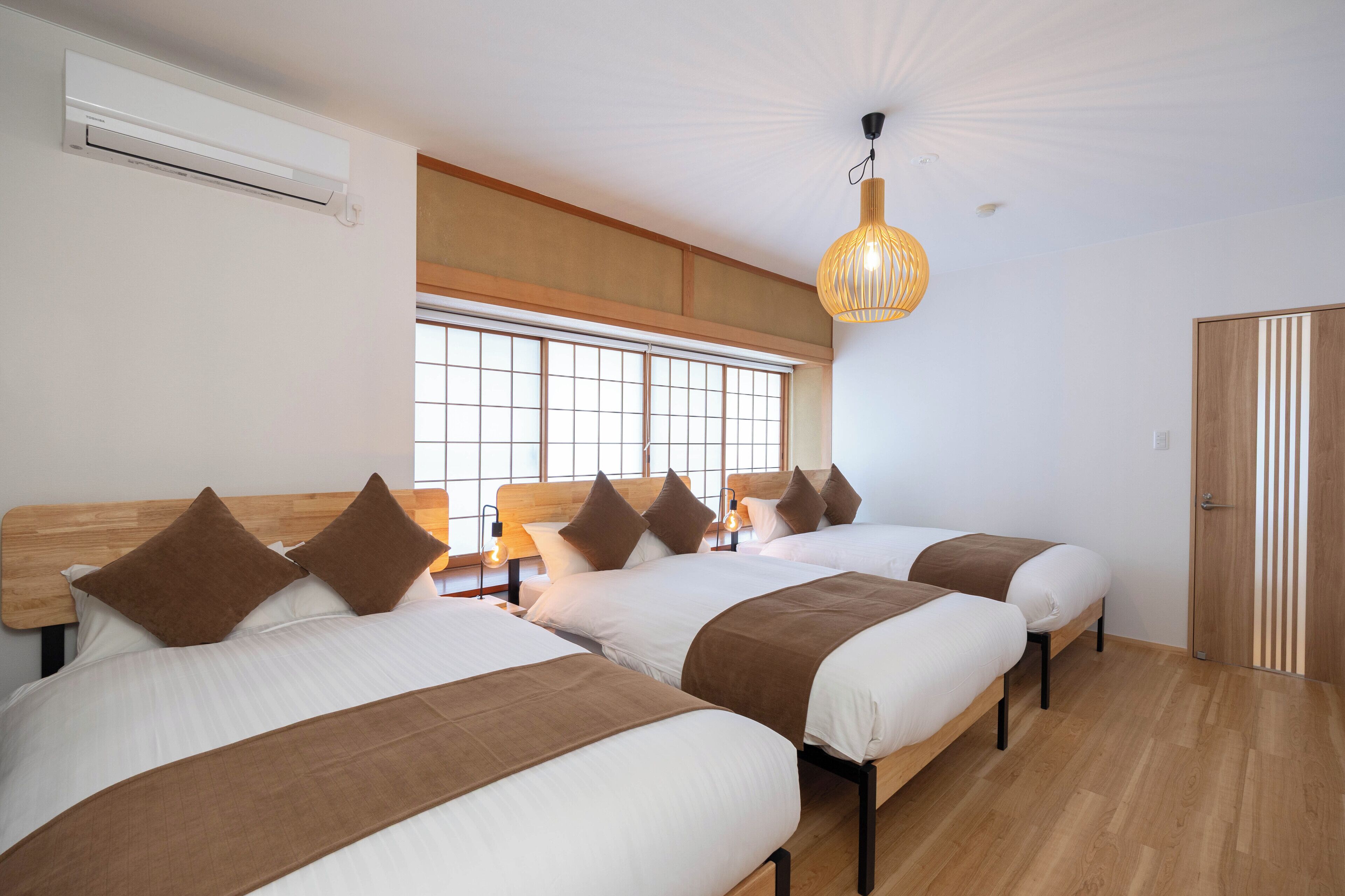 Une chambre d’hôtel avec trois lits, une grande fenêtre avec stores, un ventilateur de plafond et une armoire en bois.