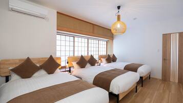 Une chambre d’hôtel avec trois lits, une grande fenêtre avec stores, un ventilateur de plafond et une armoire en bois.