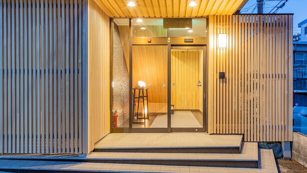 Ein moderner Gebäudeeingang mit Holzfassade, Glasüren und einer kleinen Außenbereich-Sitzgelegenheit.