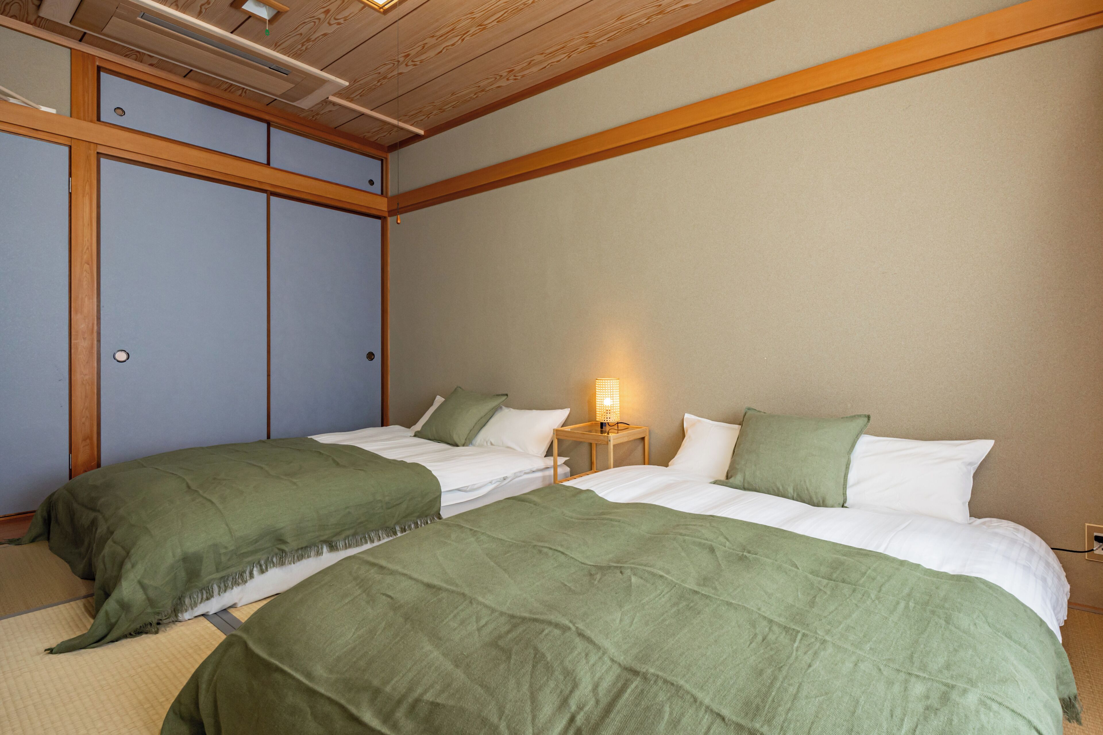 Una habitación con dos camas, cada una con ropa de cama verde y almohadas blancas. Entre las camas hay una mesita de noche con una lámpara. La habitación tiene vigas de madera en el techo y una puerta corrediza a la izquierda.