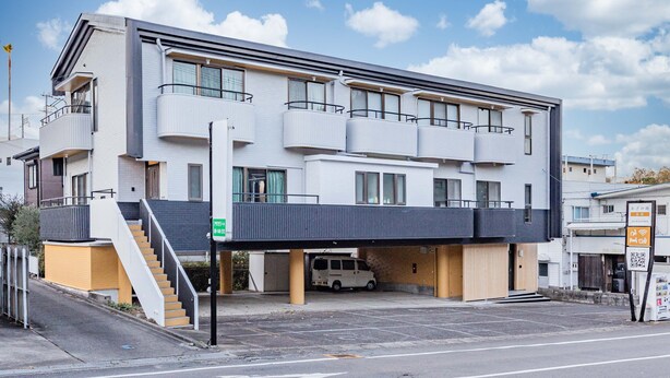 Un edificio moderno de varios pisos con un área de estacionamiento techada, una escalera que lleva a la entrada, y una furgoneta estacionada debajo.