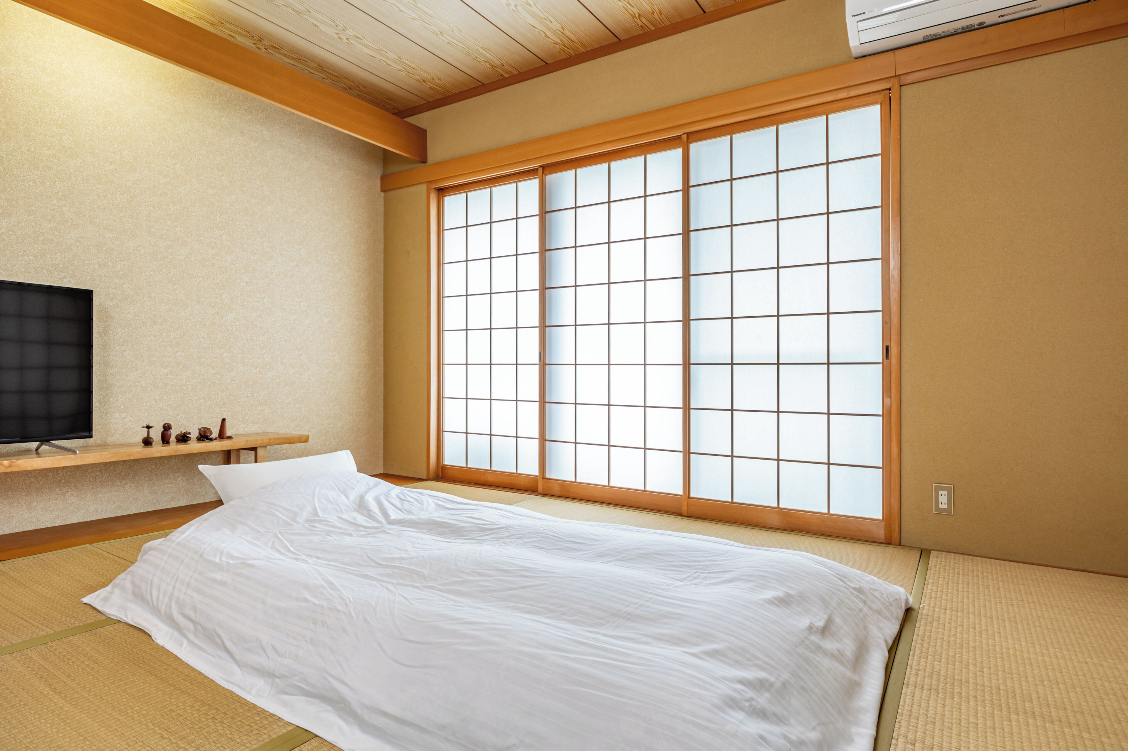 A traditional Japanese-style room with tatami flooring, a bed with white bedding, a television on a shelf, and a large shoji screen window.
