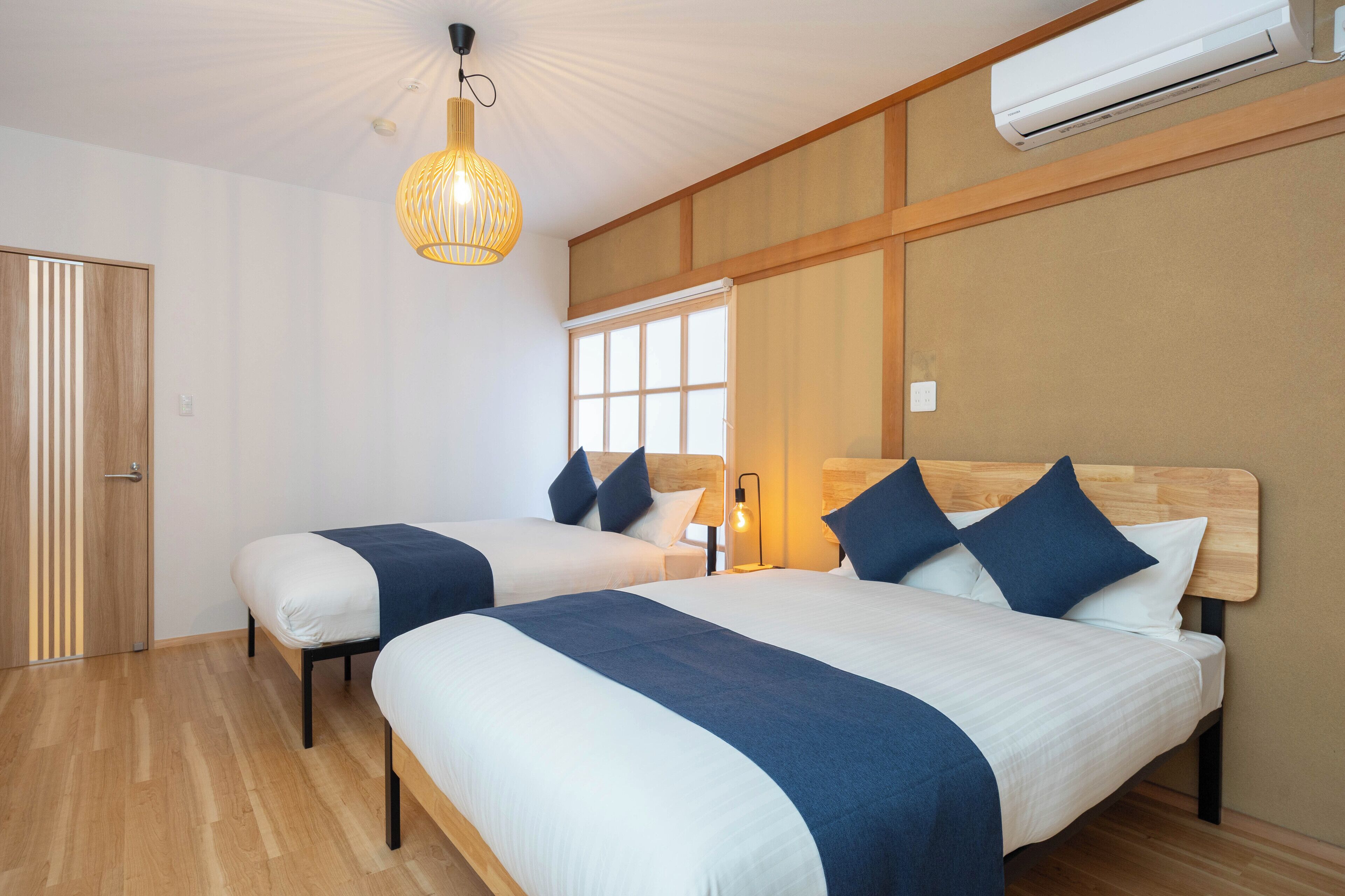 A hotel room with two beds, wooden flooring, a ceiling fan, and an air conditioning unit.