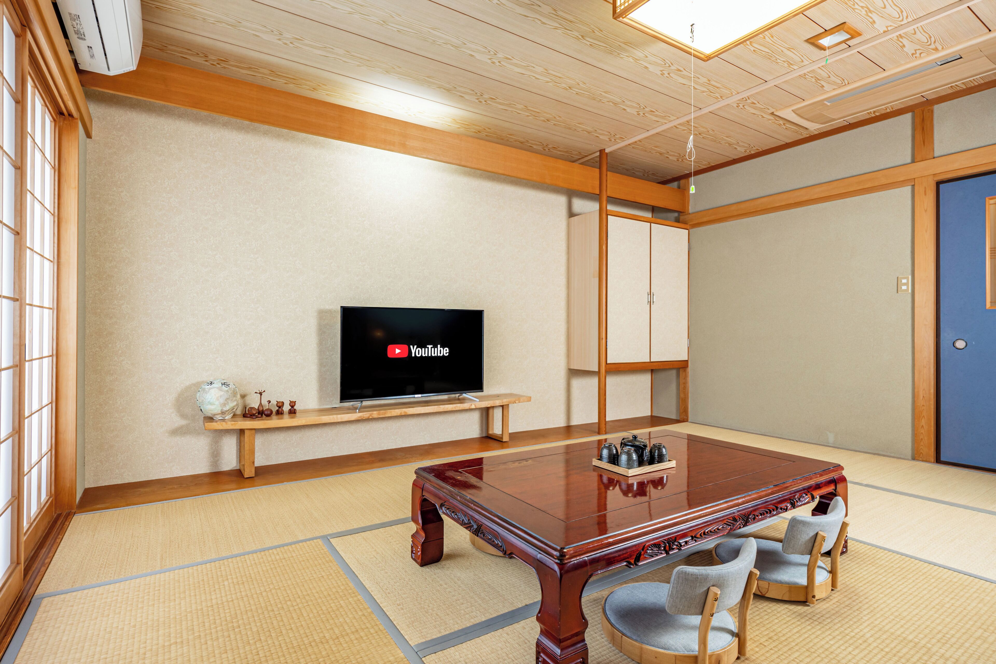 Perinteinen japanilaistyylinen huone, jossa on tatamimatto, puinen pöytä ja tuolit, televisio ja kaappi.