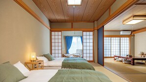 Ein traditioneller japanischer Raum mit Tatami-Bodenbelag, niedriger Holzzieldecke und Schiebetüren. Zwei Betten mit grüner Bettwäsche, ein kleiner Tisch mit Lampe und durch einen Shoji-Bildschirm ein Blick auf einen Balkon.