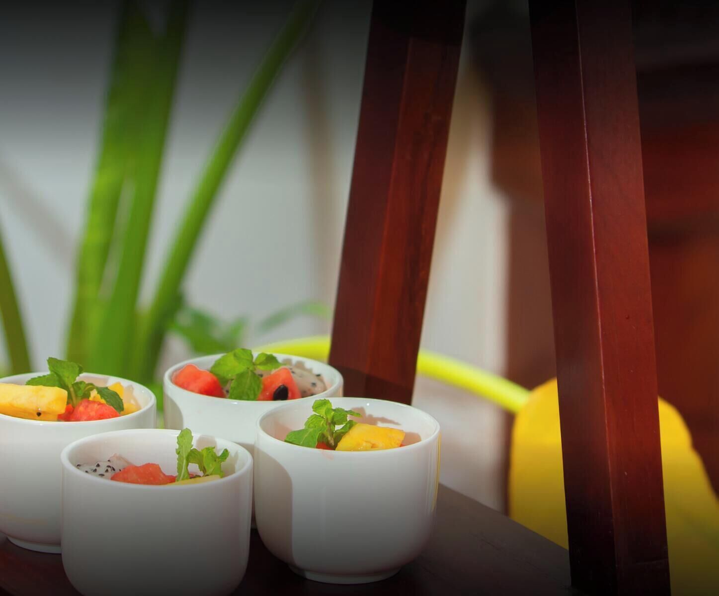 Cuatro tazones blancos con fruta y menta sobre una mesa, con una planta verde desenfocada y una silla de madera al fondo.