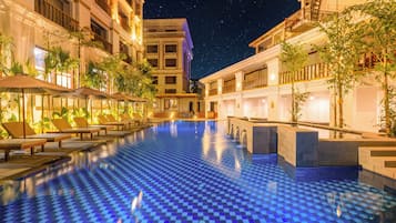 A pool area with lounge chairs and a checkered floor, surrounded by buildings with balconies and night sky.