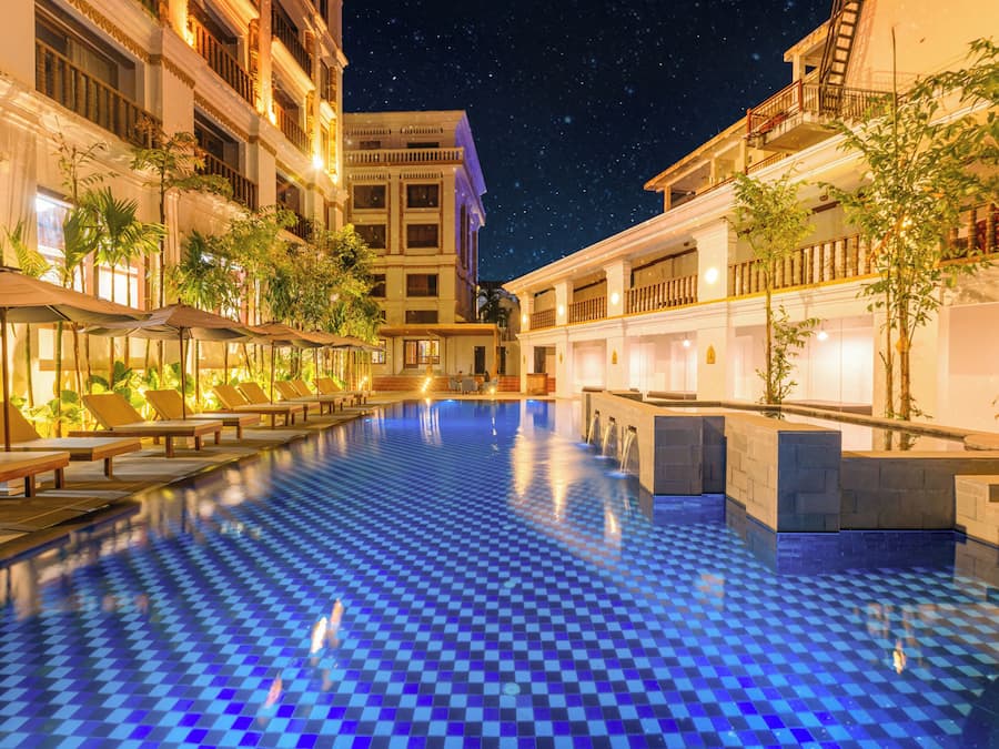 A pool area with lounge chairs and a checkered floor, surrounded by buildings with balconies and night sky.
