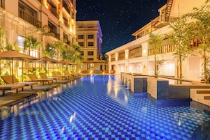 Área de piscina com espreguiçadeiras e piso quadriculado, cercada por edifícios com varandas e céu noturno.